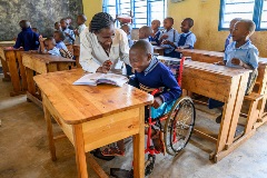 A laughing boy sits in his wheelchair with a smiling teacher bending down next to him in a classroom full of children in Rwanda.