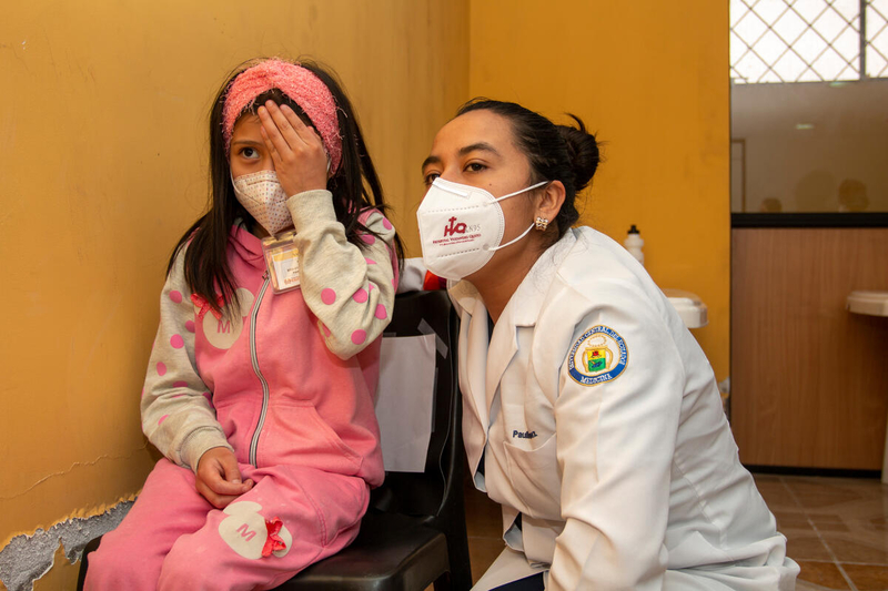 A girl sitting on a chair reading an eye chart with one hand over her eye while a health worker bends down next to her.