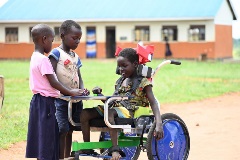 A girl with a physical disability in Uganda sits in a wheelchair and plays a game with two other children outside a classroom.